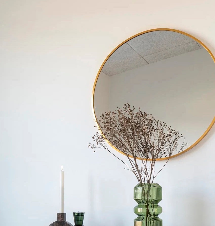 Rund spegel i mässing upphängd på vägg med en grön vas under och en torkad kvist